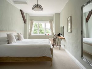 Calm and elegant bedroom at Bampton Longhouse in Devon with soft green tones, a large mirror, and a bright window overlooking the gardens.